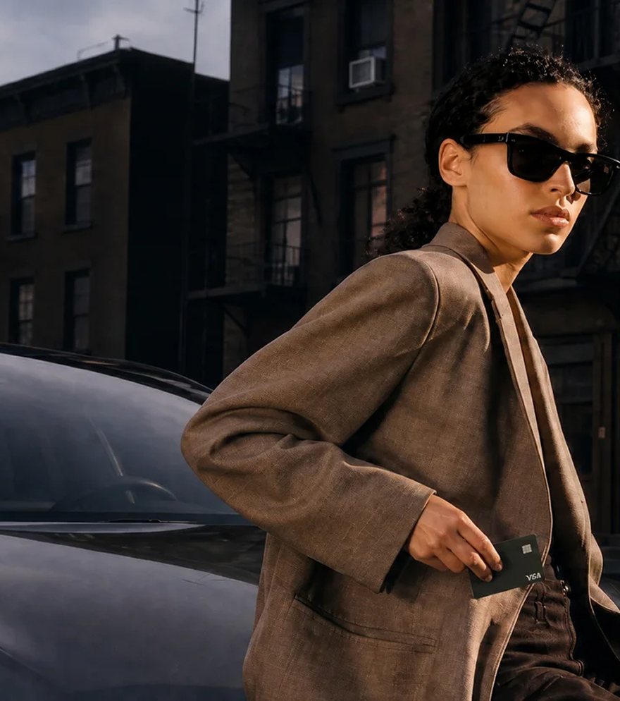 A woman in sunglasses walking past a parked car on a city street, holding a payment card