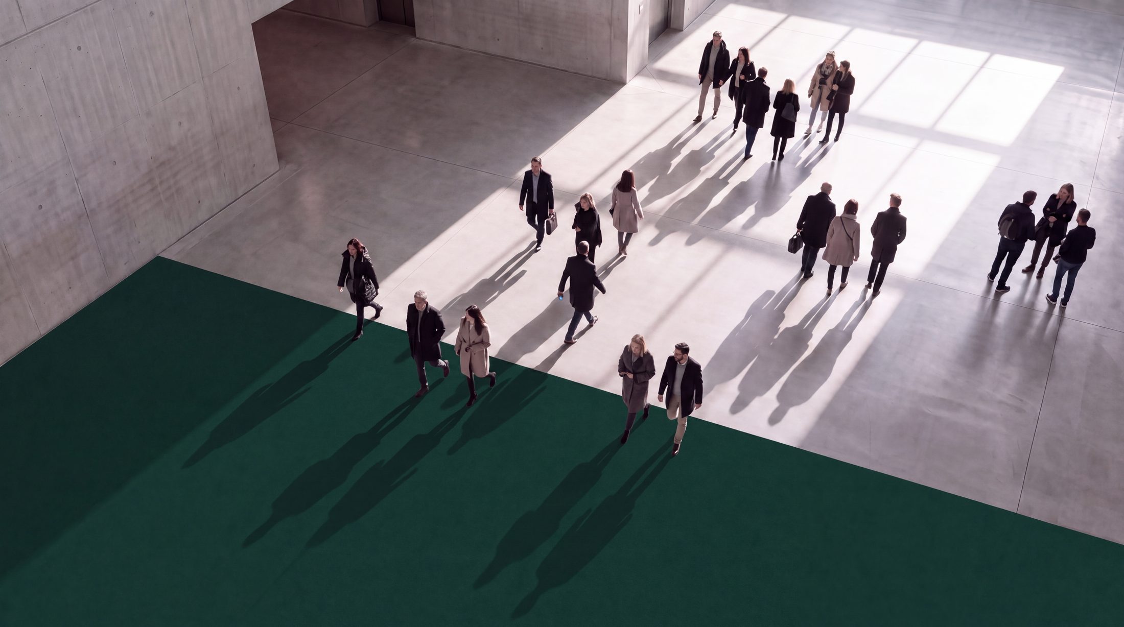 Commuters crossing a concrete plaza, long shadows falling across a deep green band of floor